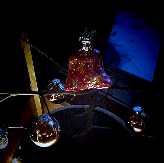 A pink and gold glass Buddha hung on a bare tree,at night. Fairy lights illuminate it and glass balls hanging nearby.