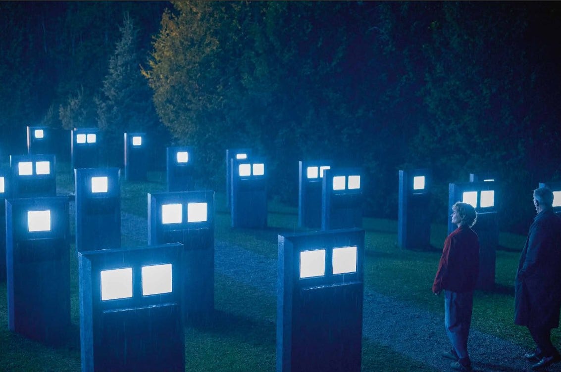 Movie still from The Shrouds. A man and woman stand outside at night, with multiple electronic screens on short pillars lining the path.