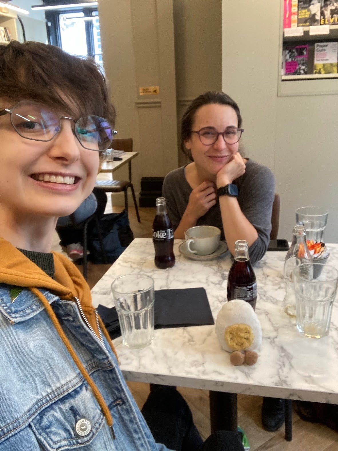 Photo of Beck and Michaela, a smiling white woman with glasses, in a cafe with a table full of empty cups and two freshly opened glass bottles of coke. Among the clutter is dippy, the soft toy egg, happy to be in the frey