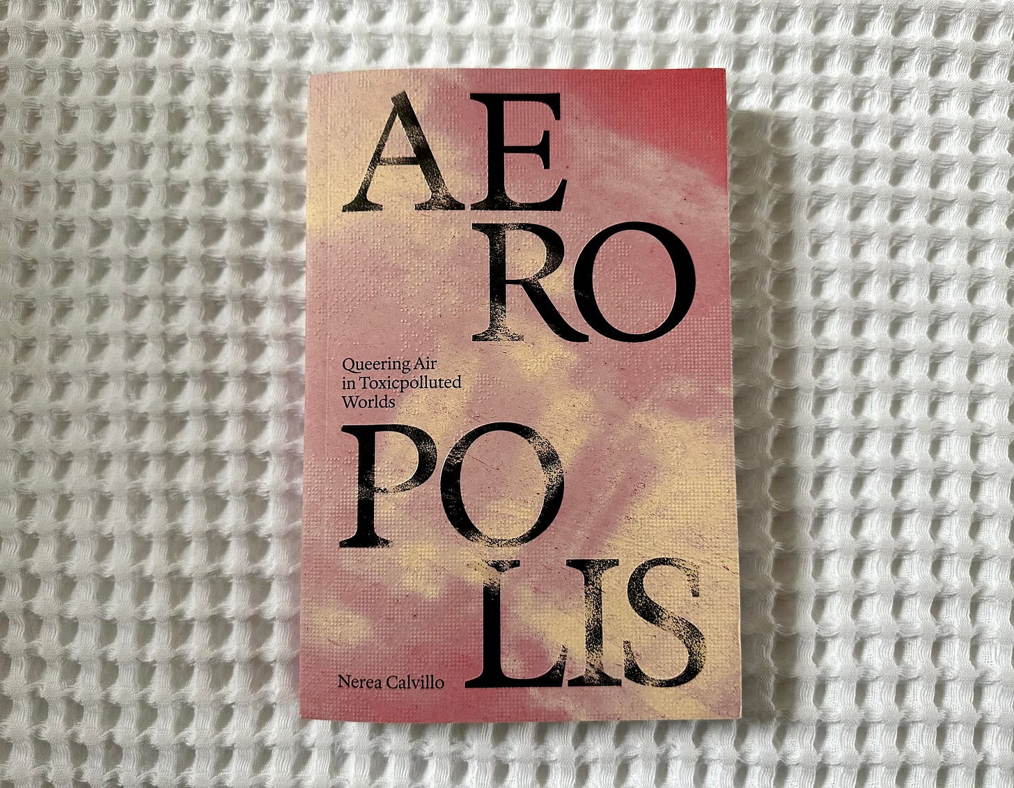 Picture of a book titled "Aeropolis: Queering Air in Toxicpolluted Worlds", by Nerea Calvillo, on a white textured blanket. The cover of the book is pink with off-white pal clouds with AE, RO, PO, LIS appearing in large capital letters on different levels across the cover.