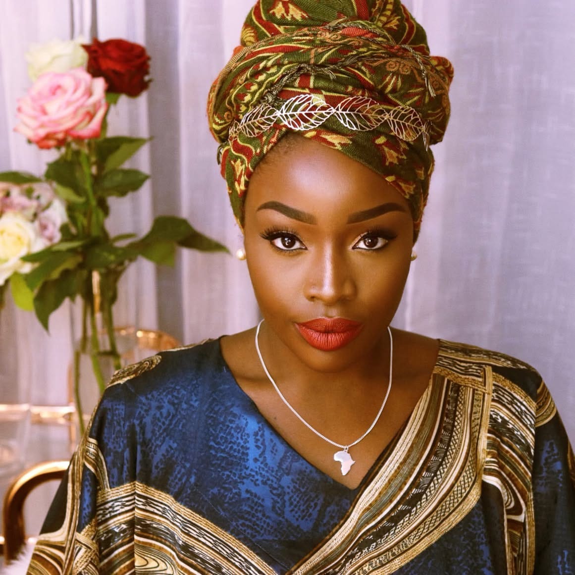 Lovette Jallow, a Black autistic author and speaker, wearing a green patterned African headwrap and a gold-trimmed blue garment. She sits confidently in front of a bouquet of roses, facing the camera with a calm, powerful gaze.