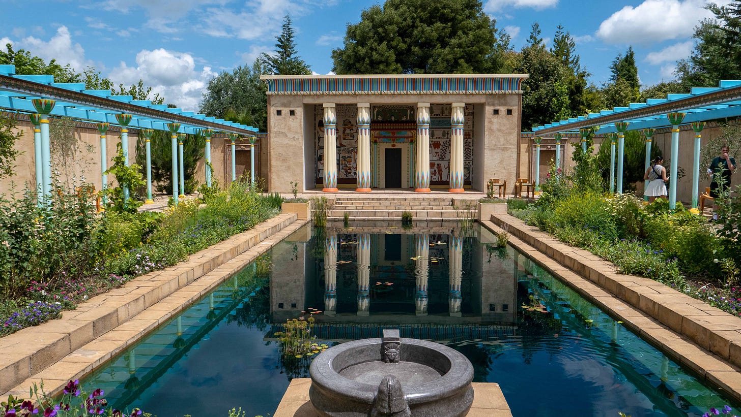 An artificially blue rectangular pool flanked by colourful colonnades leads to a highly decorated area under a roof held up with Egyptian style columns.