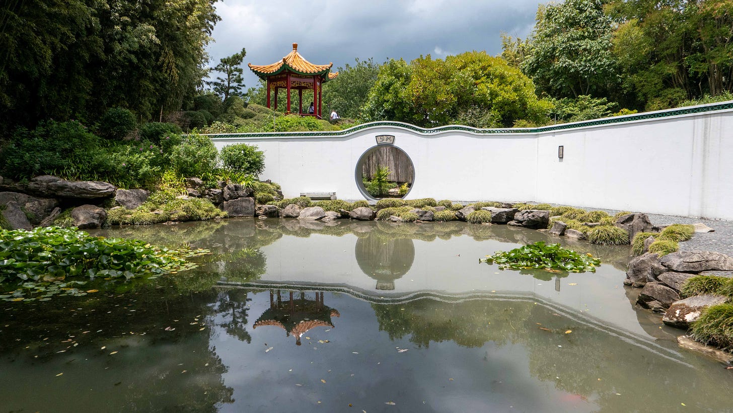 A pool reflecting a stormy sky, pagoda on a rise and the moon gate in a white wall.