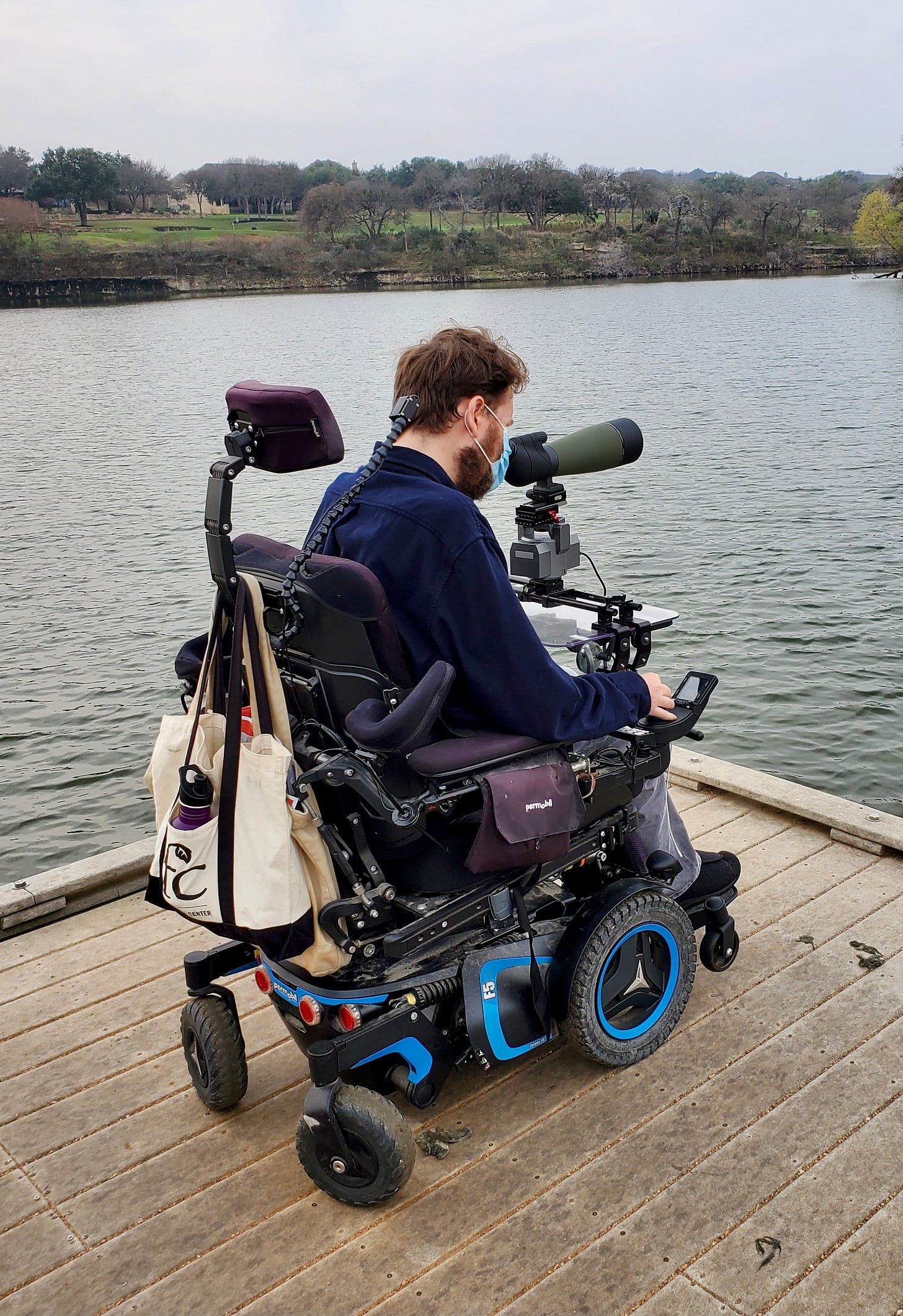 Eric birds from the end of a dock on an artificial lake. Seated in his large power wheelchair, he leans his head forward to gaze through a spotting scope mounted to his wheelchair tray. The opposite shore is rocky, with lawns and leafless trees beyond. The water is as gray as the sky.