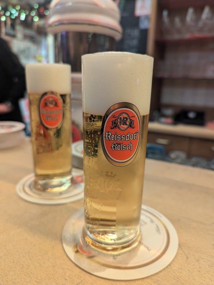 Two glasses of Kölsch with the gold and red Reissdorf Kölsch logo printed on them.