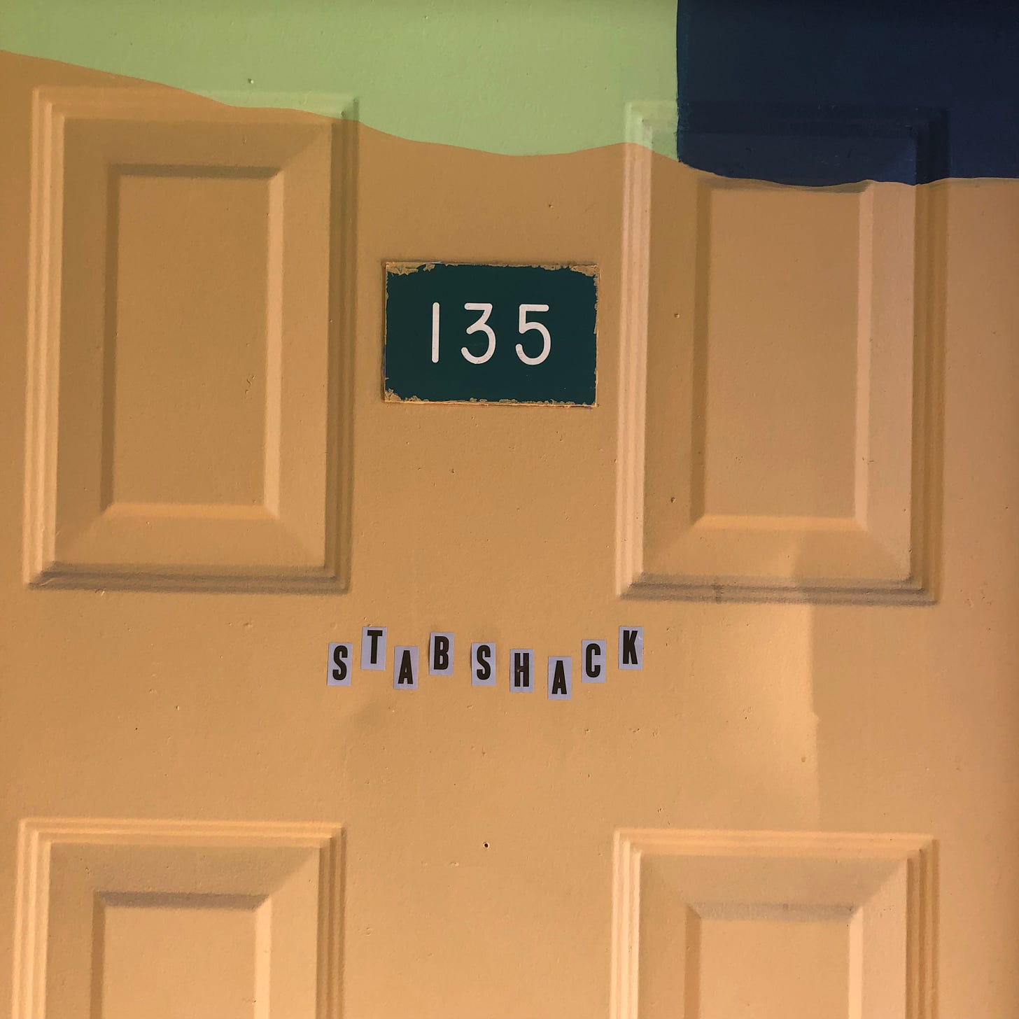ID: yellow door with mint green and navy blue patches a white on green sign sits at the center number 135 below it in lettering the title stabshack is displayed across the door.