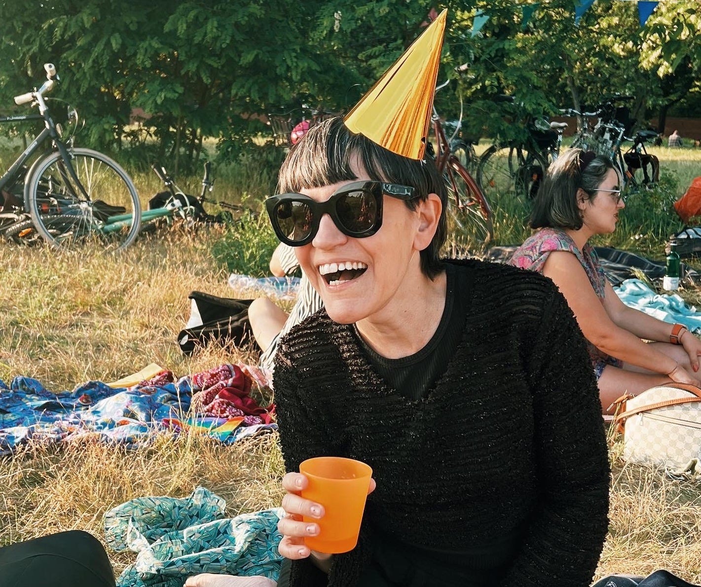 Imagem: Mulher cabelo curto,franja, oculos escuros grandes, rindo e com um chapéu em cone dourado. Segura um copo e está sentada na grama. Atrás há outras pessoas.