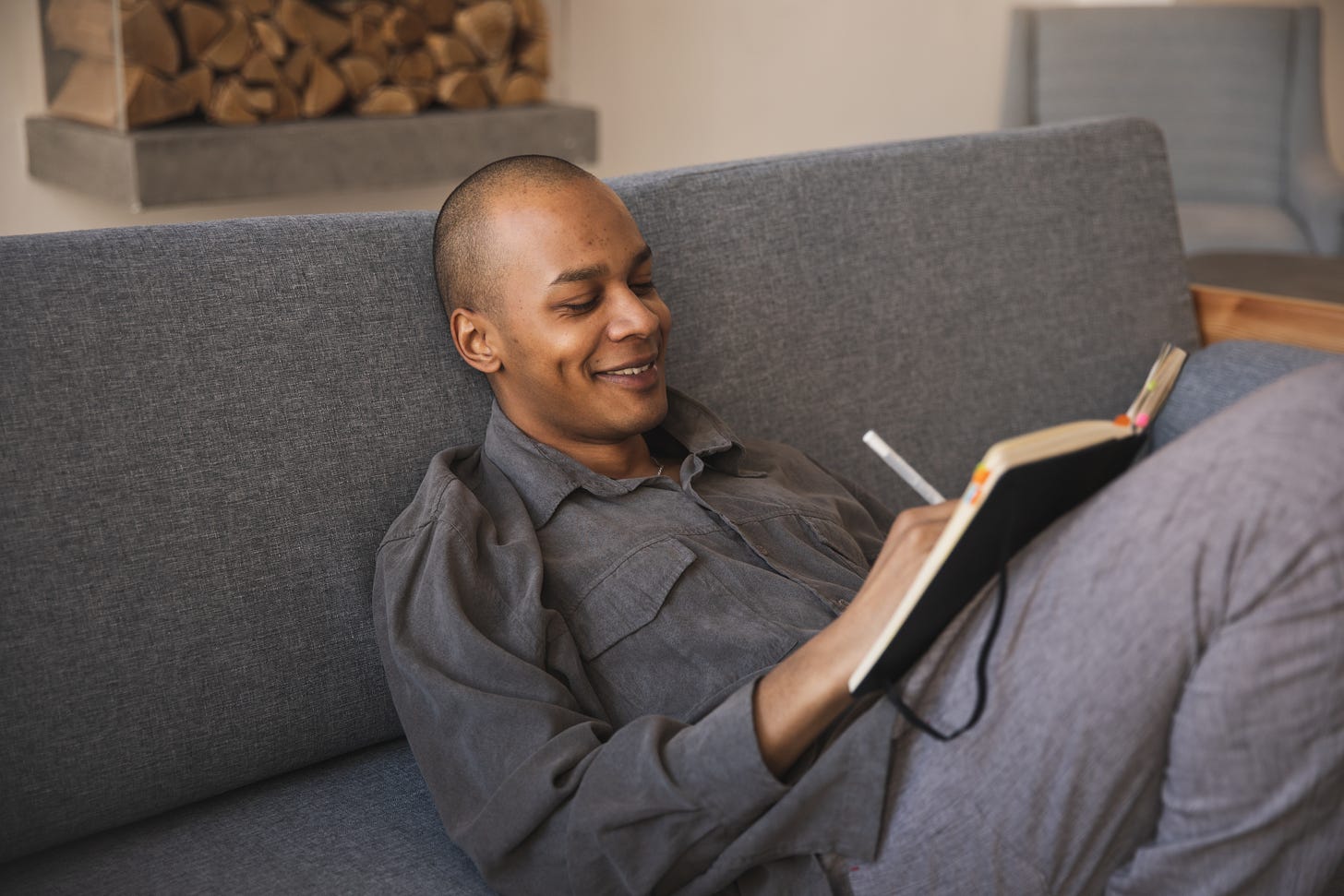 a man in a grey shirt reclines on a grey sofa writing by hand into a notebook and smiles