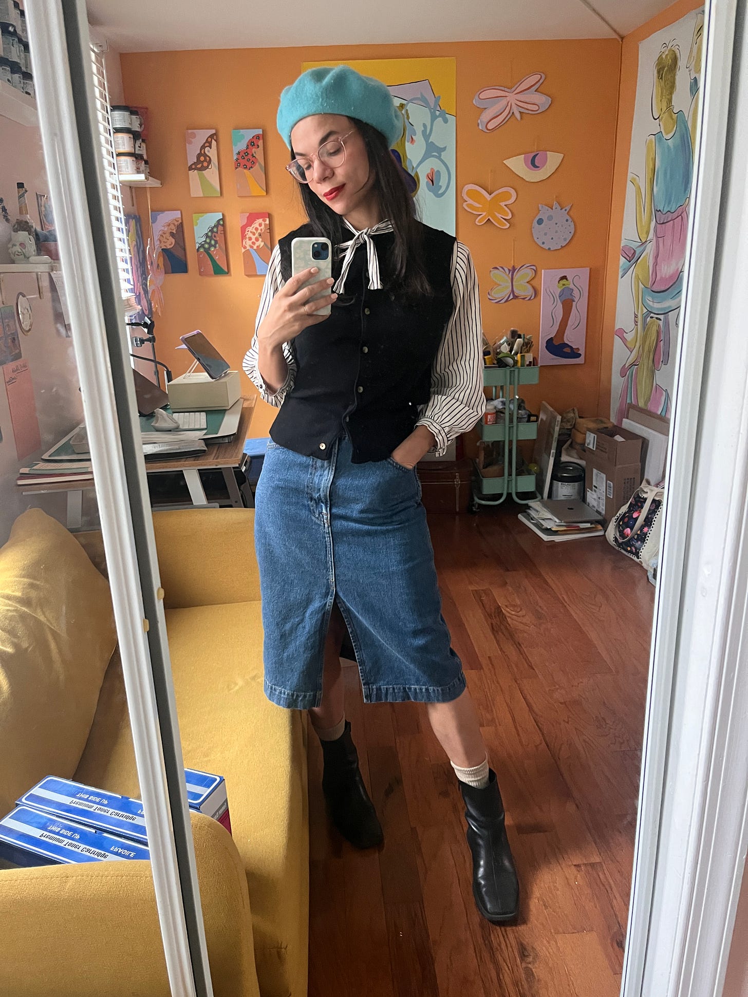 The artist Cindy Lozito stands in front of her studio mirror wearing a robin's egg blue beret, a black sleeveless button vest, a vintage 1950s pinstriped button-down with a bow, a midi denim skirt, and black ankle boots with beige socks peeking out.