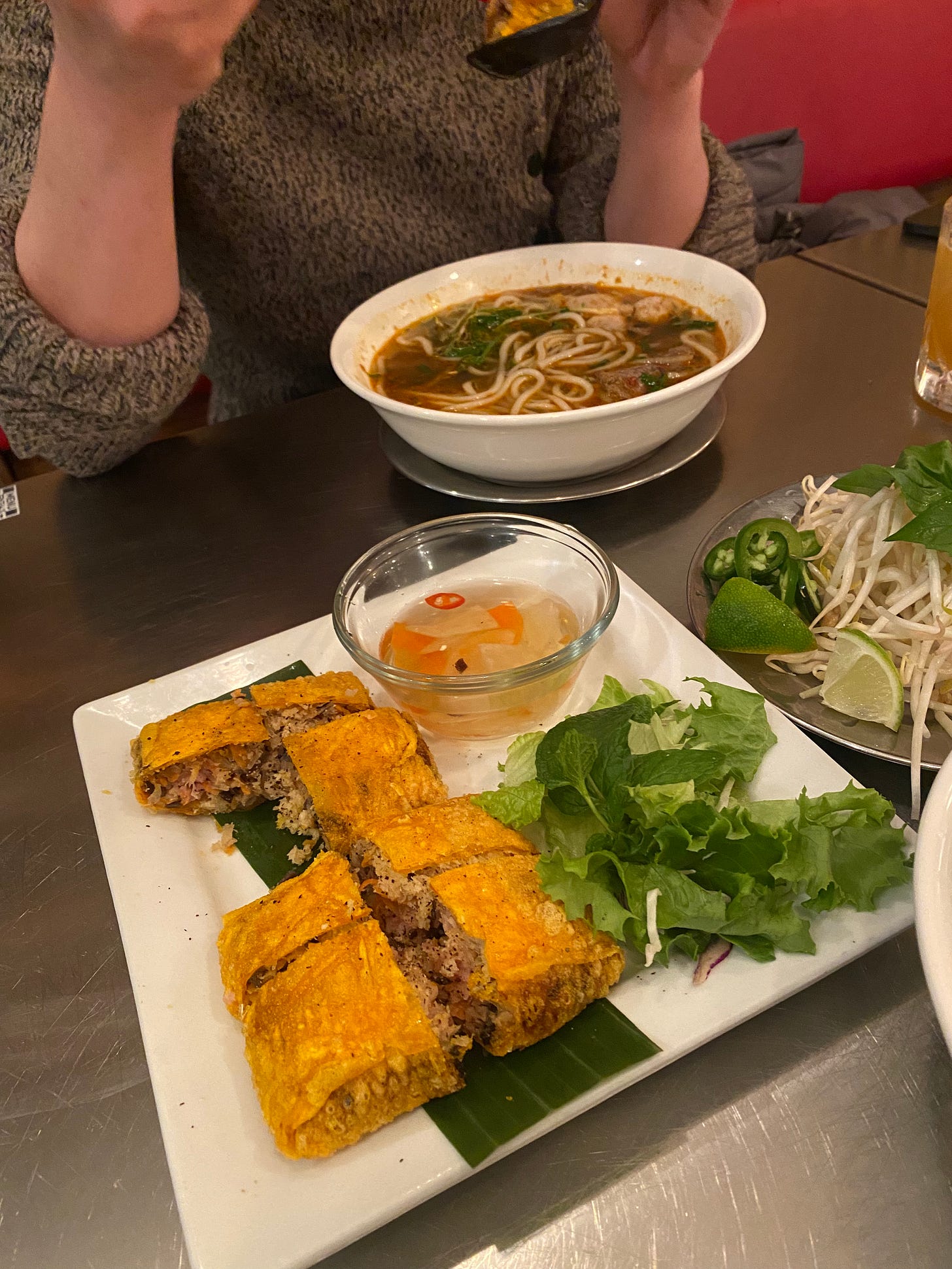 The crab pocket described above, a yellow-orange fried rice paper filled with meat and vegetables next to a small salad and a dish of clear fish broth. Next to the plate is a plate of pho garnish, bean sprouts and herbs and lime and jalapeño. Liang is eating a bowl of spicy pho in the background.