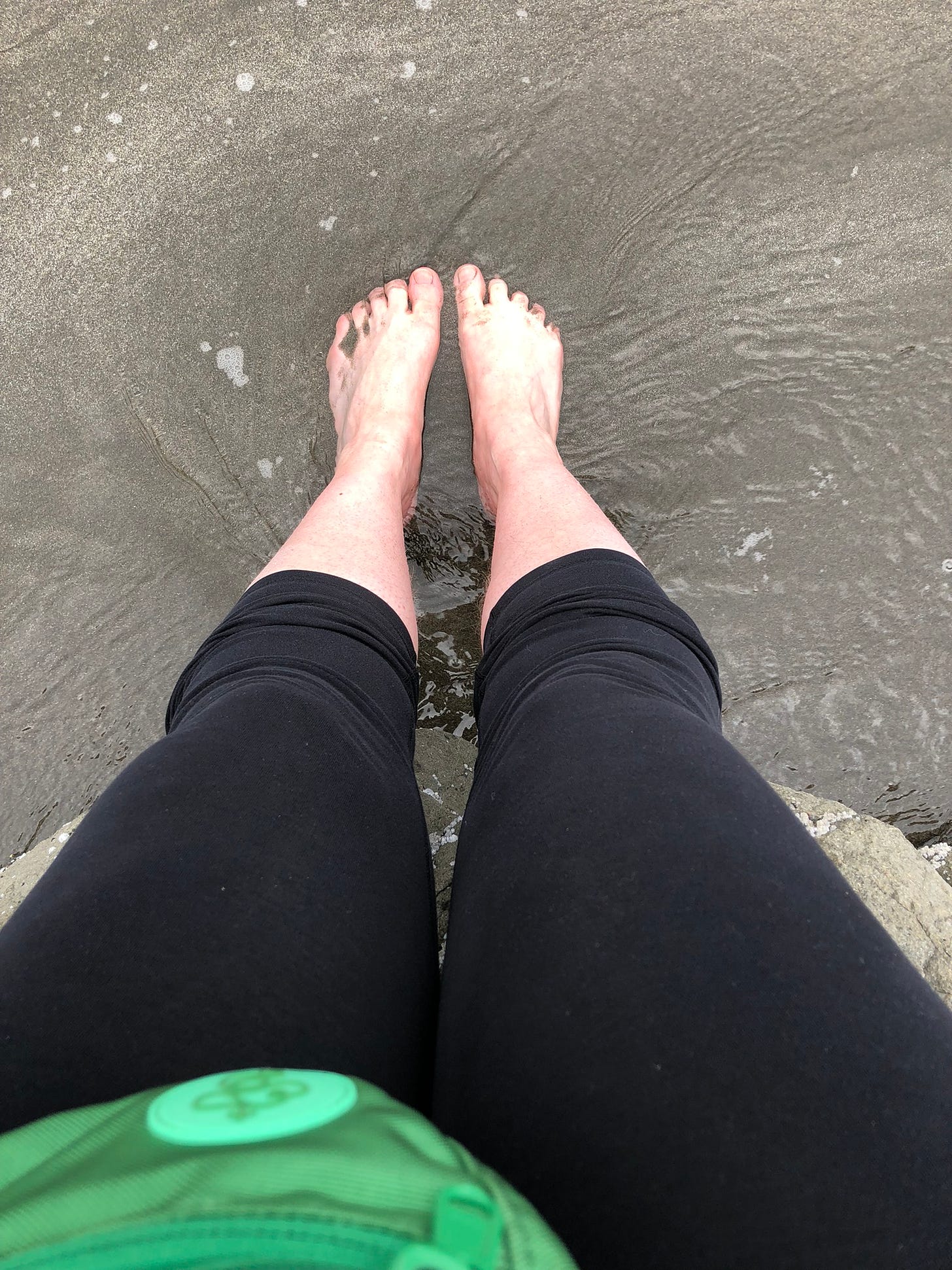 from a pov looking down, the edge of a green fanny pack above two legs with black leggings scrunched upward with bare feet resting on the sand where a wave has just receeded