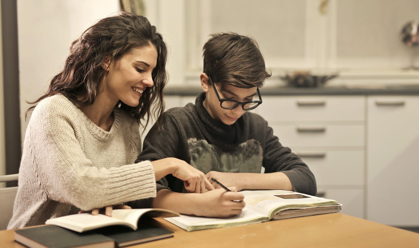 To siblings work on homework together. The older, a woman, is pointing out something to her younger brother.