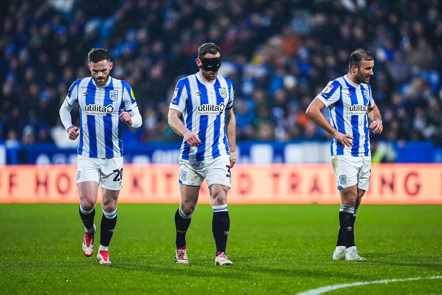 Huddersfield Town players Ollie Turton, Tom Lees (wearing a black protective face mask), and Herbie Kane stand on the pitch at the John Smith’s Stadium during the Reading game. They are wearing the 2024/25 home kit, featuring blue and white striped shirts, white shorts, and black socks. Raindrops are visible in the background. Photograph credit: Huddersfield Town.