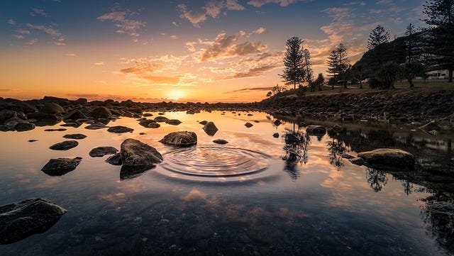 Sunrise over a rocky pool of water, with tree-lined hills to the east. The pool of water contains a few ripples as if a pebble were just tossed in.