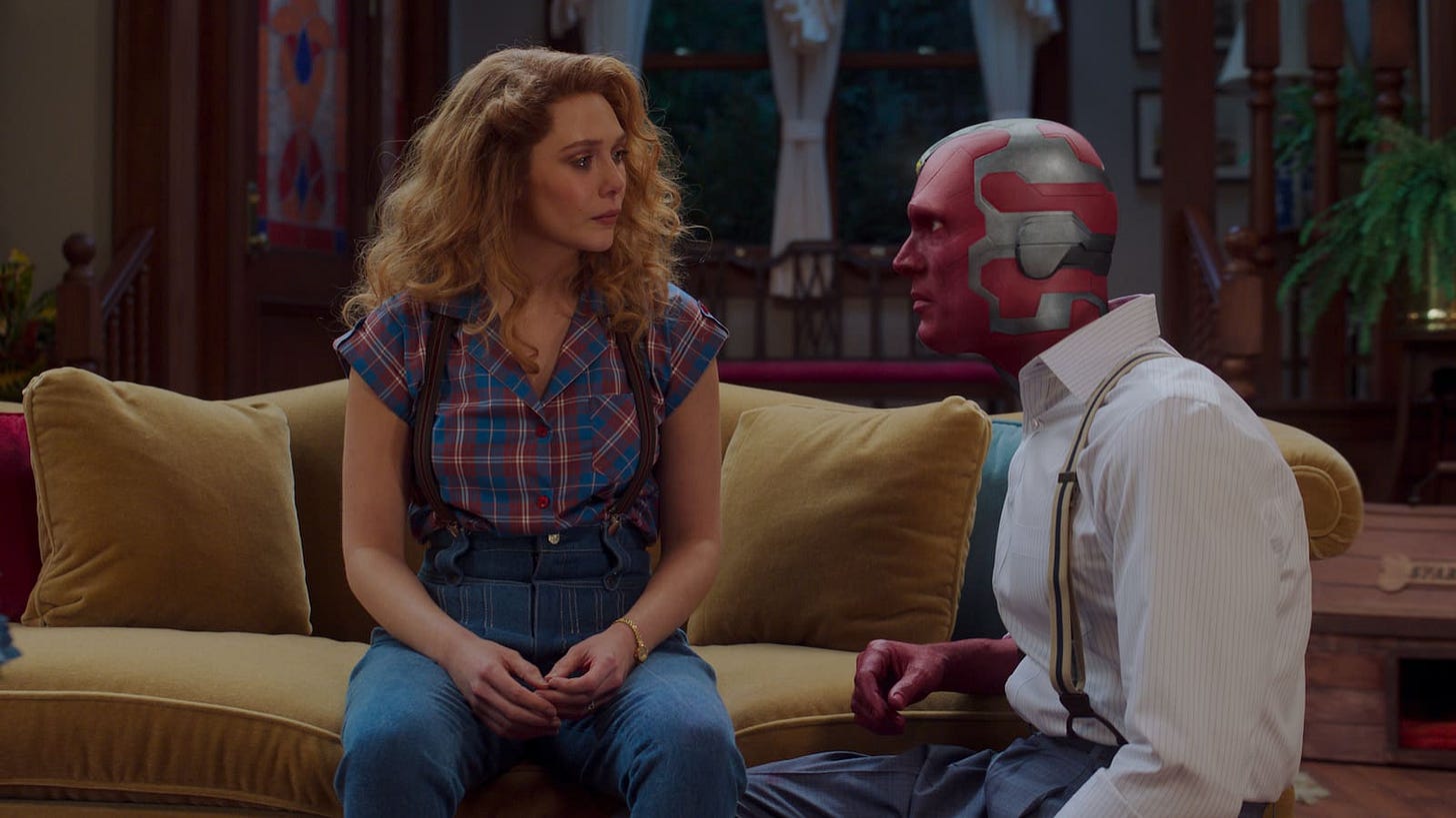 Wanda sits on yellow couch in tears as Vision kneels beside her in his work clothes.