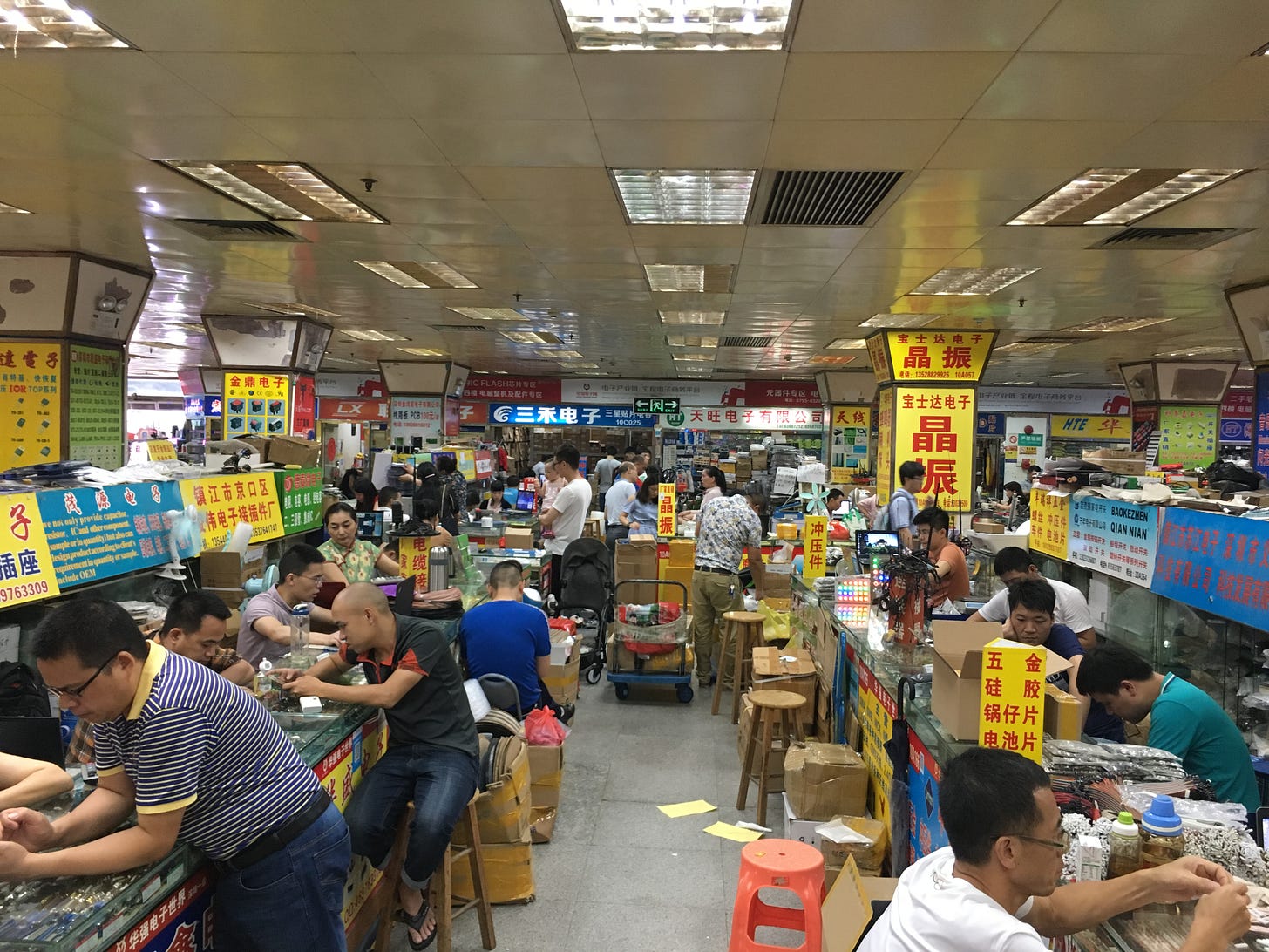 Crowded electronics market with colorful signs, booth with people sitting and plastic stools