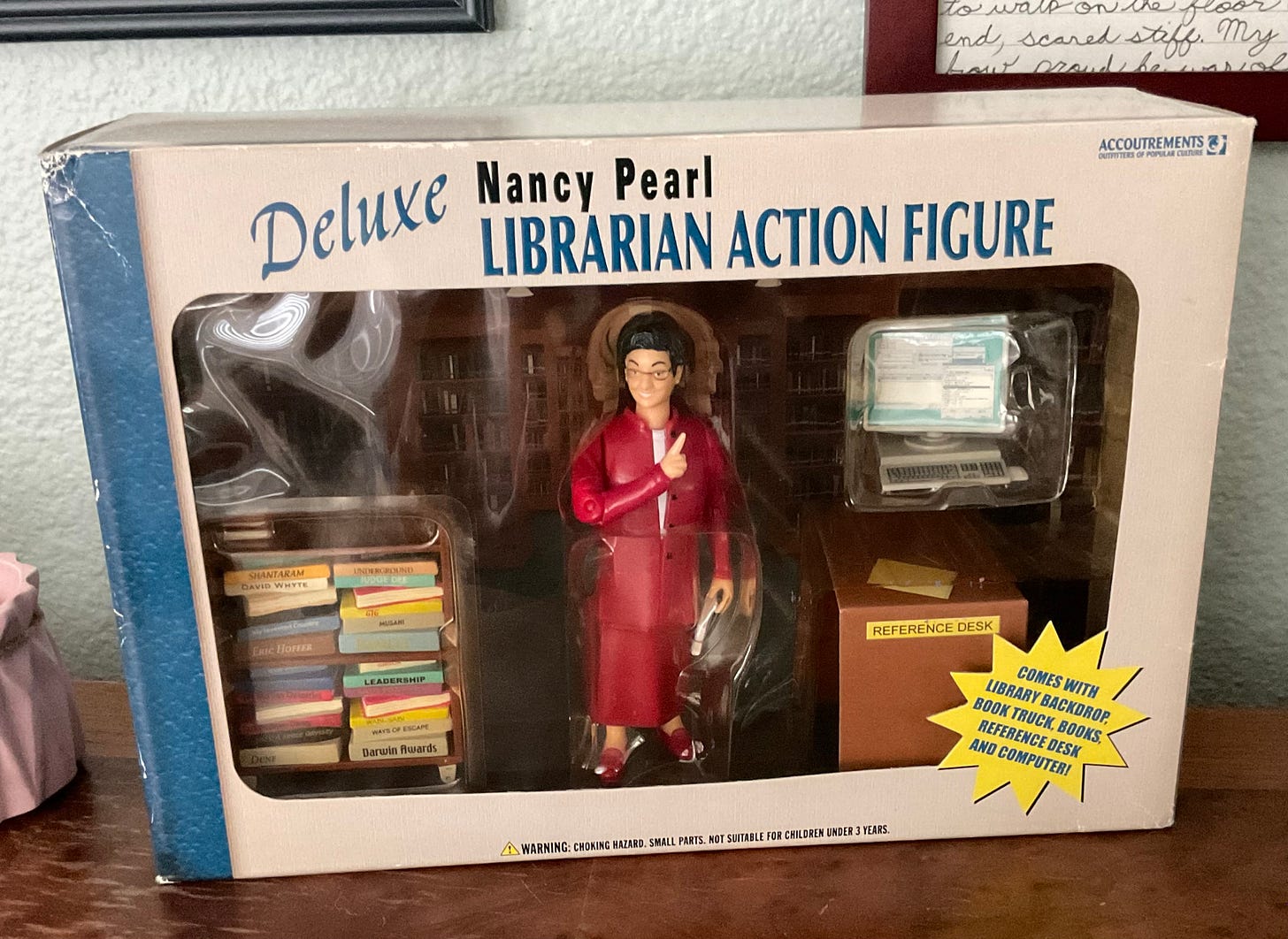 A librarian action figure with accessories. The librarian is wearing a red suit. The accessories include a desk, a cart full of books, a computer, and a library backdrop. 