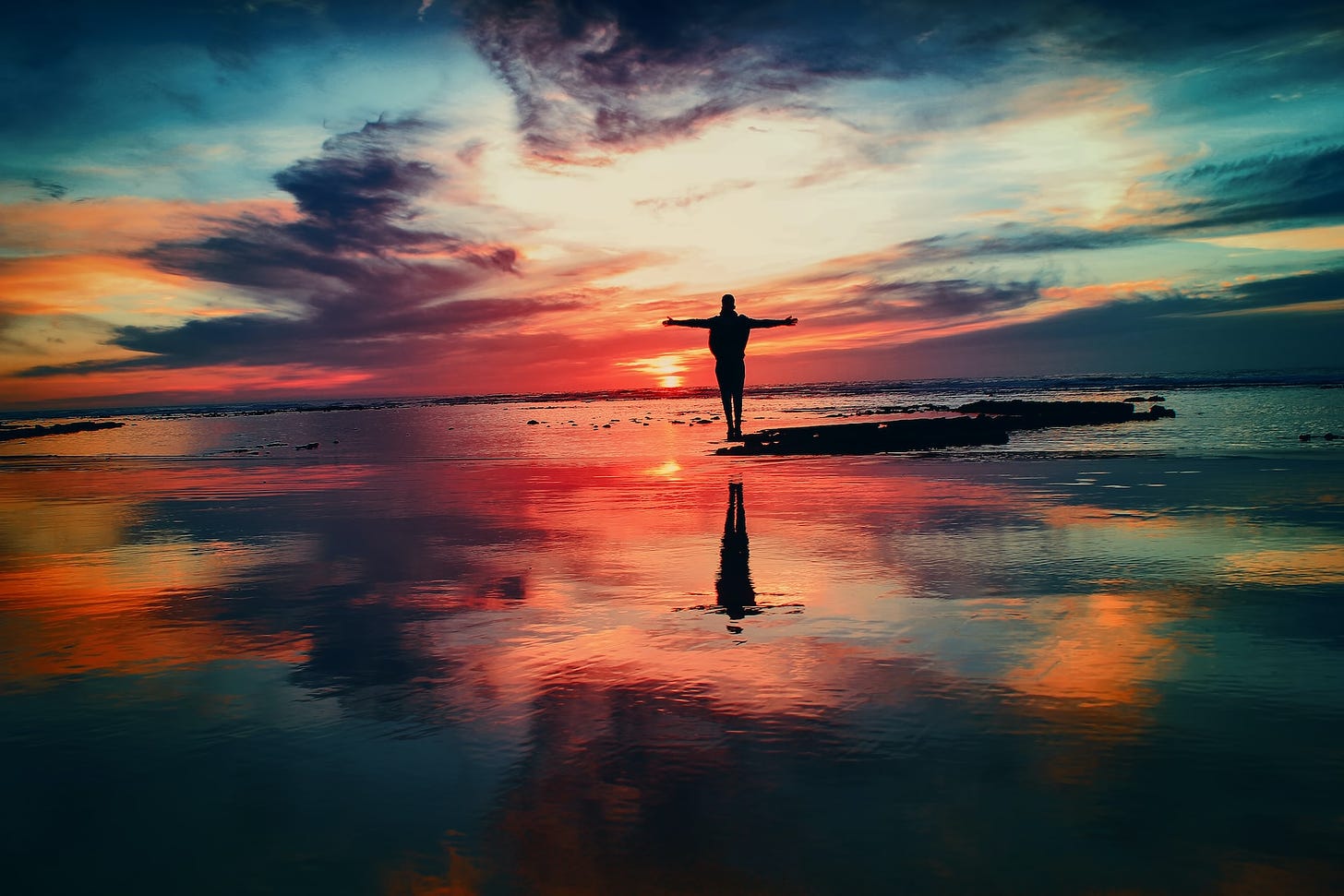 Person on a rock in a reflective body of water with a sunrise or sunset in the background.