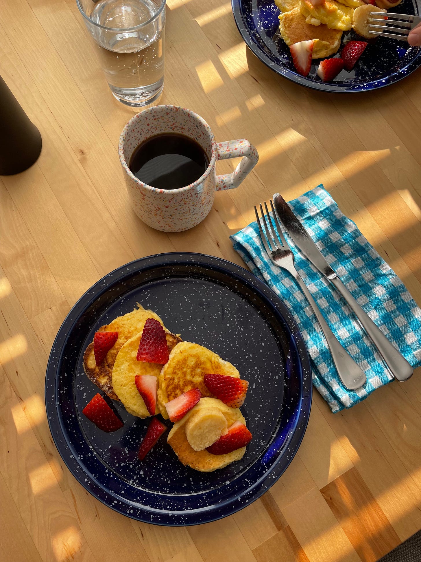 Speckled plate with four mini ricotta hotcakes, sliced strawberries sprinkled on top. A slice of honeycomb butter is on the plate. There is a cup of filter coffee on the table and a blue and white gingham napkin.