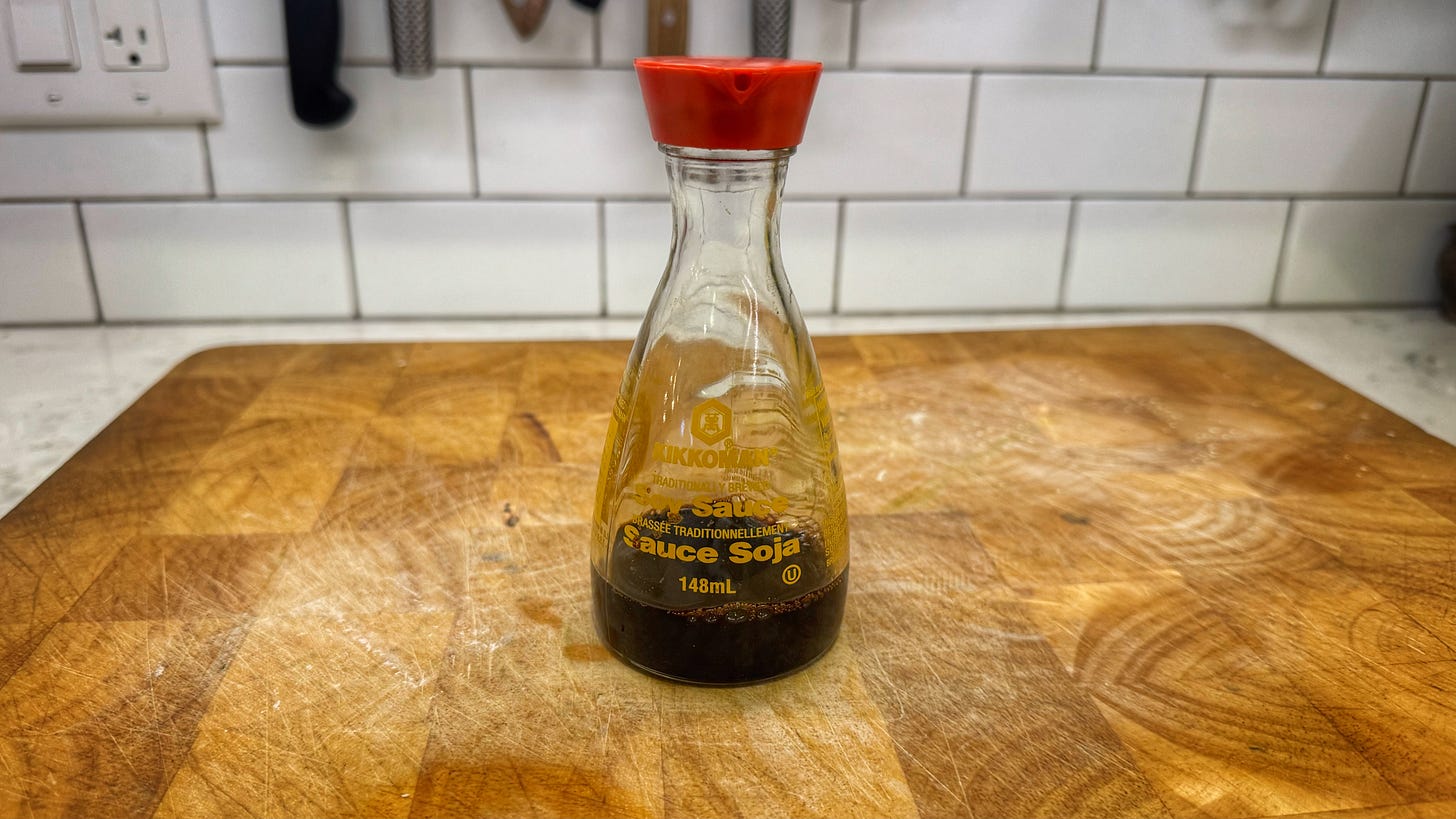 An almost empty, glass Kikkoman soy sauce dispenser sitting on top of a wood cutting board.