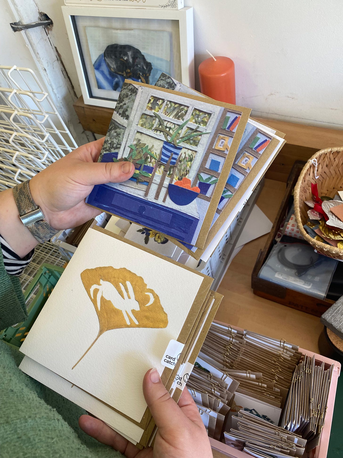 Two hands hold a stack of greetings cards made from original art pieces by Megan Fatharly, one a collaged room scene with plants in front of a window and the other a gold printed gingko leaf with a white silhouette of a have and crescent moon in.