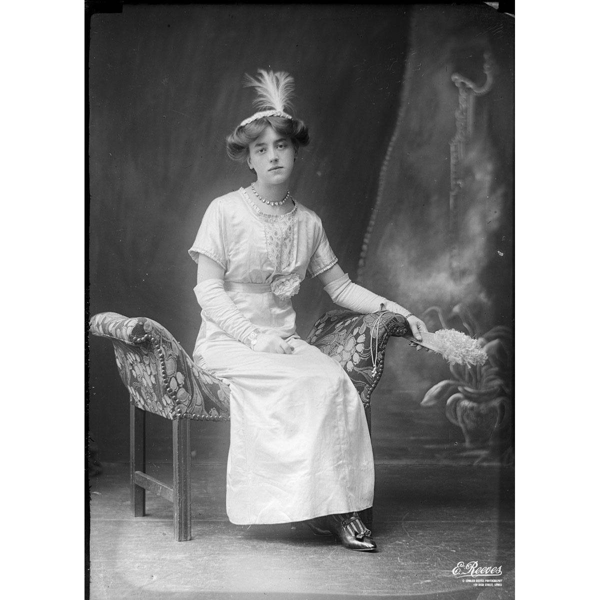 An old black and white photo of a well dressed woman sitting on a chair in a photography studio