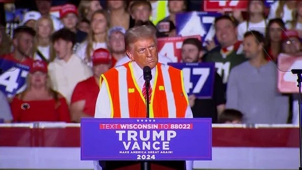 A person wearing an orange vest and a white and blue vest standing at a podium with a sign and a crowd of people behind him

Description automatically generated