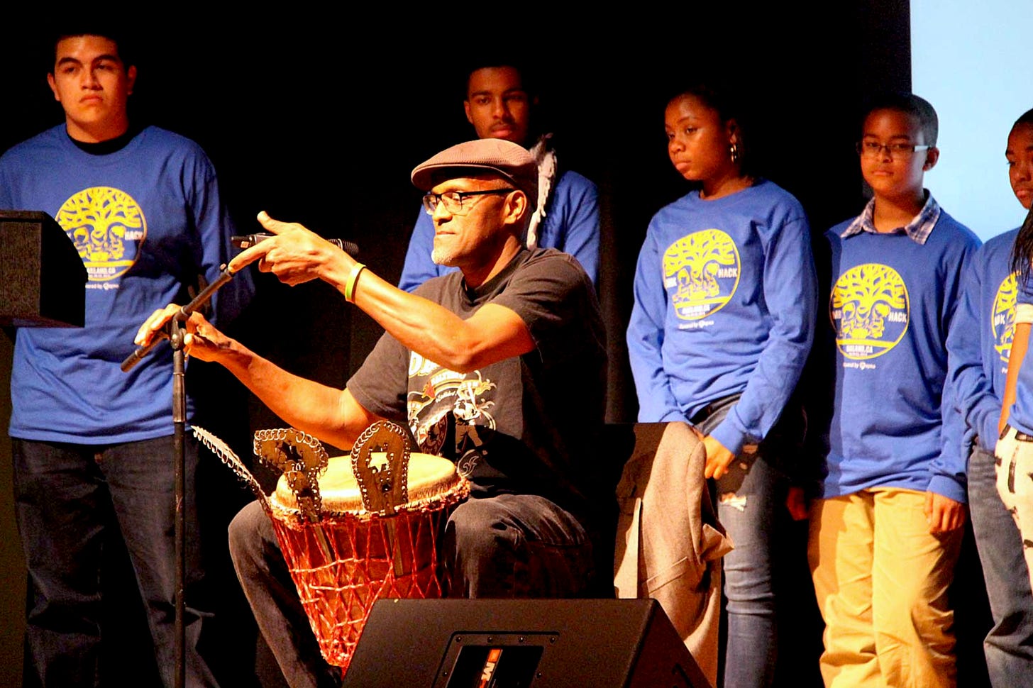 Greg seated with an African drum, children and youth stand behind him in matching blue shirts with oaktree design