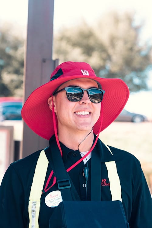 Free Young employee smiling in a red hat outdoors on a sunny day, wearing sunglasses and uniform. Stock Photo