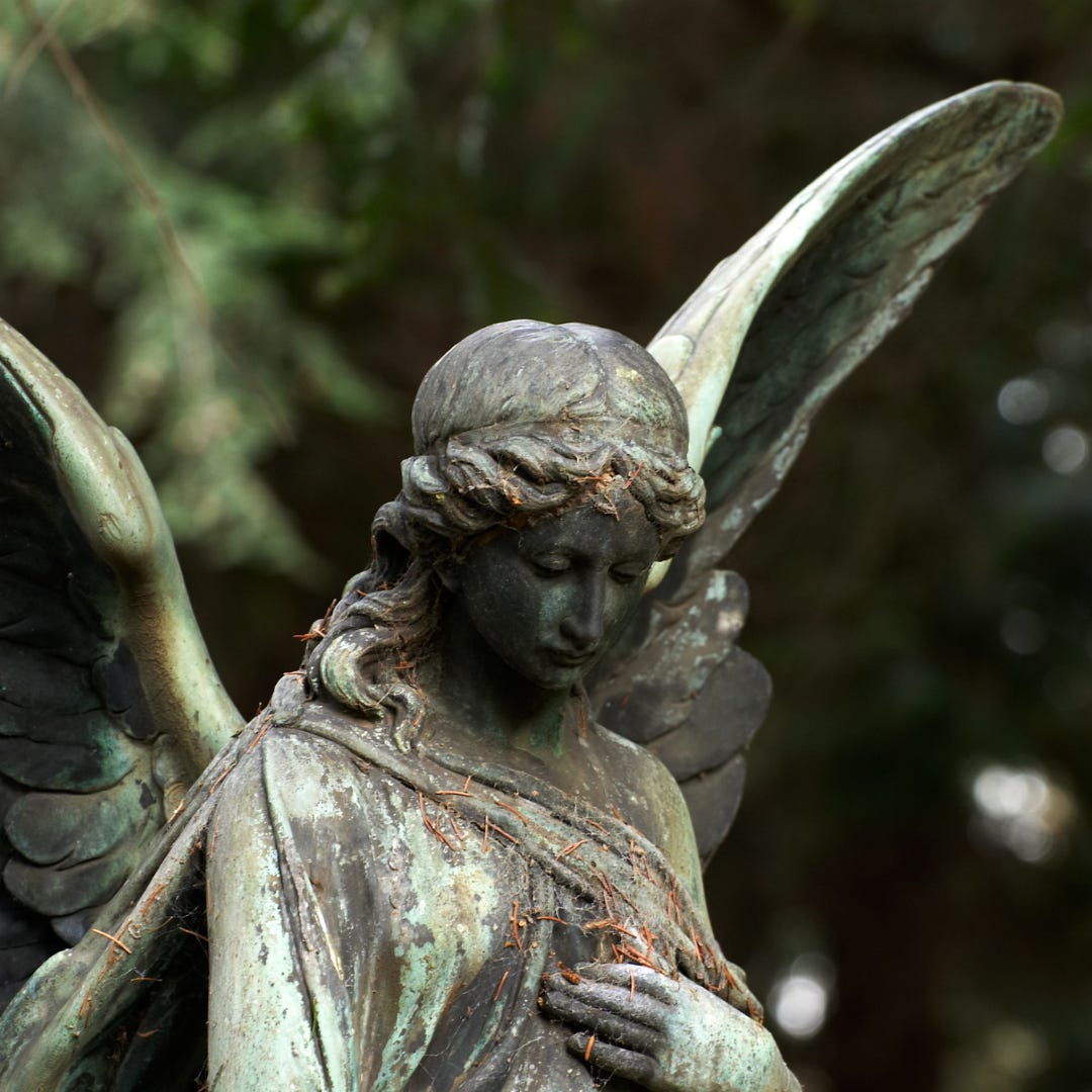 A statue of an angel with a weathered green patina gazes toward the earth with one hand on their heart.