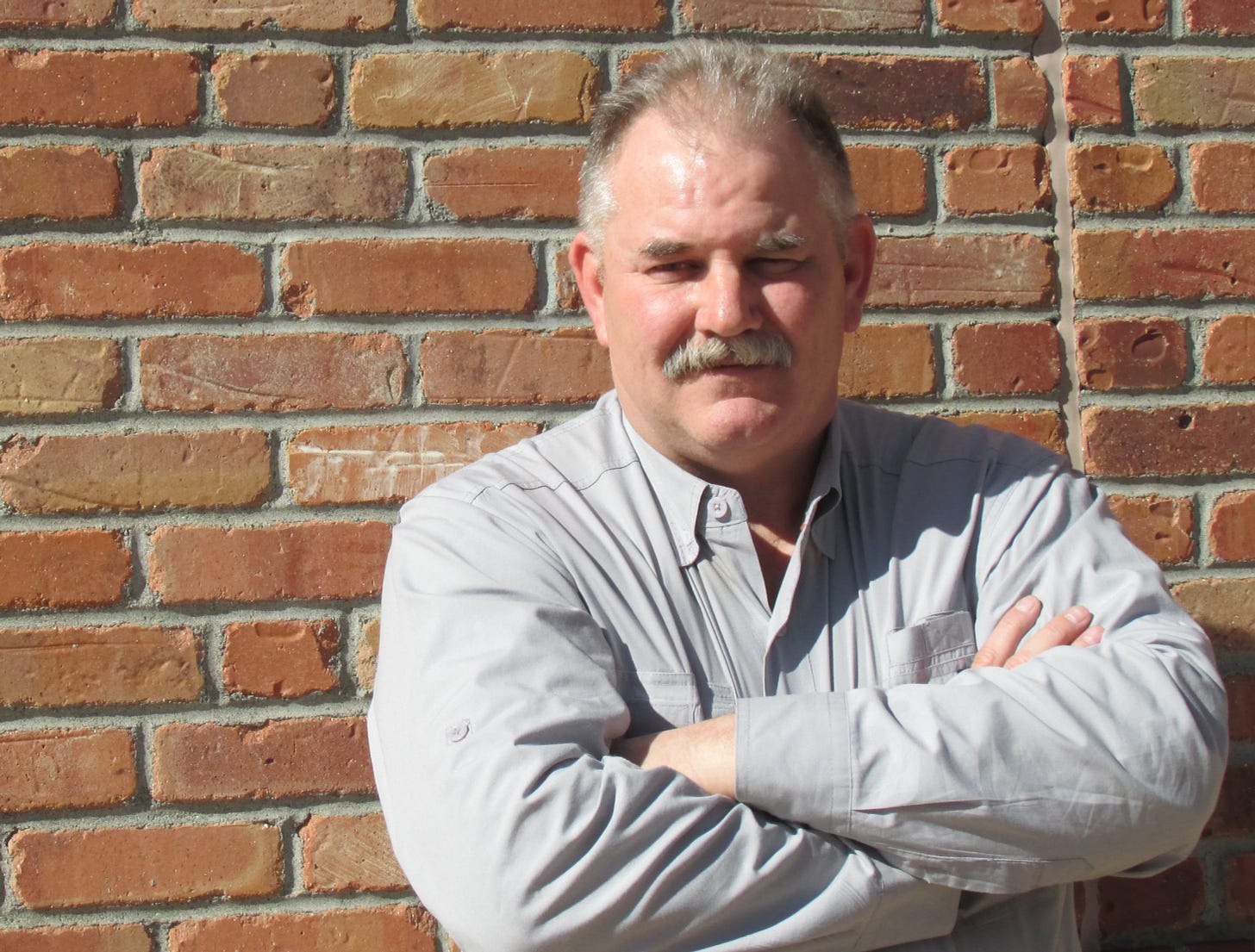 man in gray work shirt with arms folded against an exterior brick wall