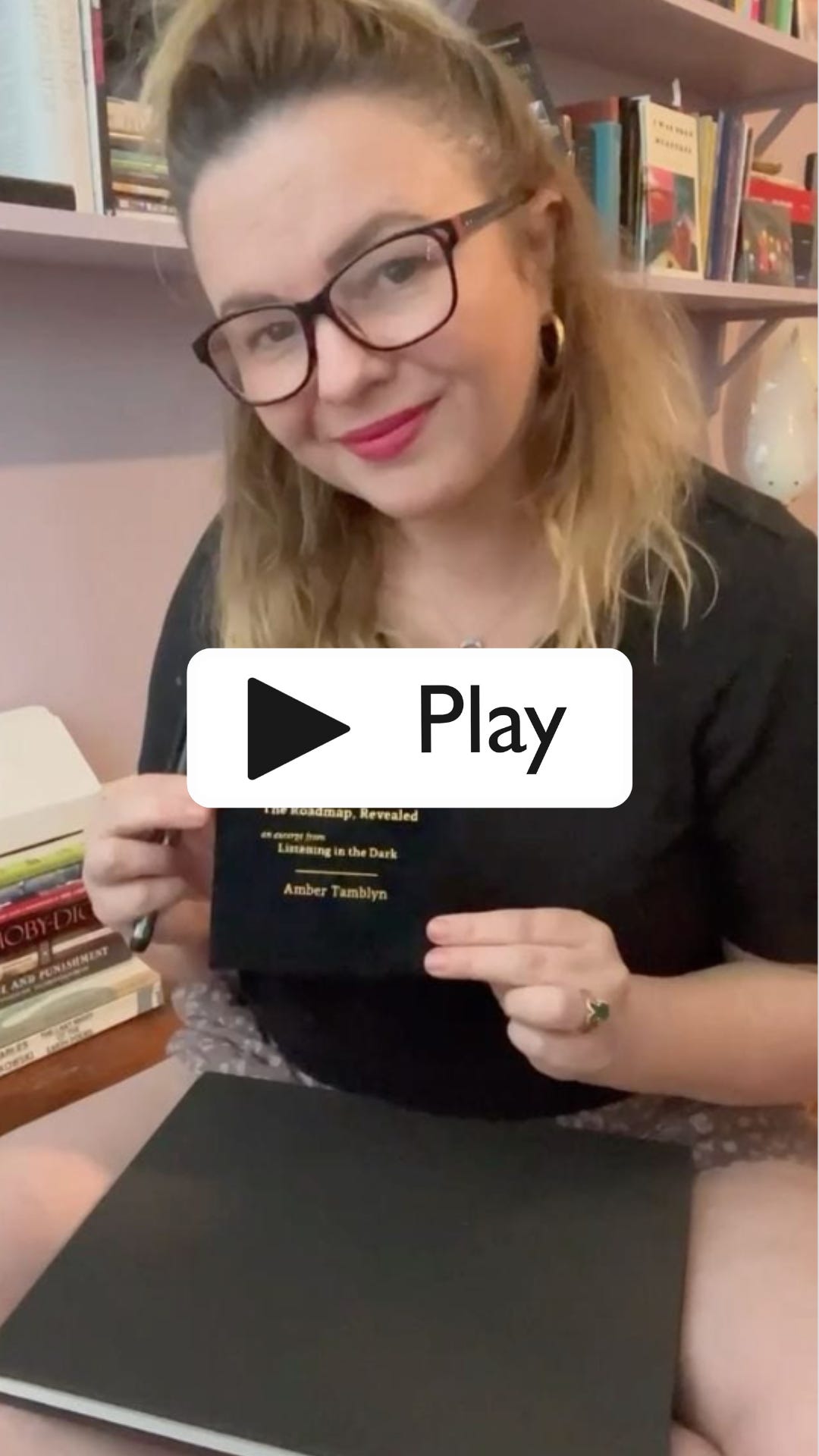 A Play button over a still from a video. Amber holds a small black book and smiles into camera.