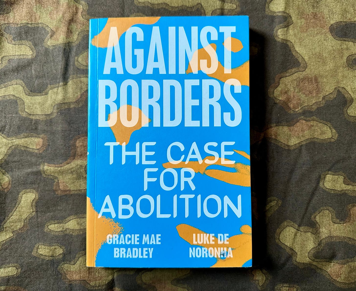 Image of the book "Against Borders" by Gracie Mae Bradley and Luke De Noronha on a dark green camouflage fabric. The cover of the book is bright blue with the title taking up most of the cover in large capital letters.