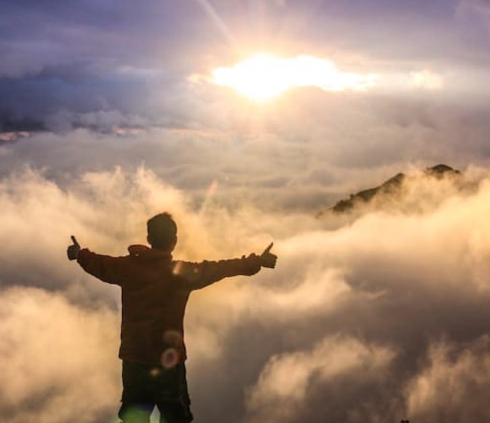 Silhouette of a young man seen from behind with arms spread, giving thumbs up as he floats in the sky amidst the clouds and the sun.