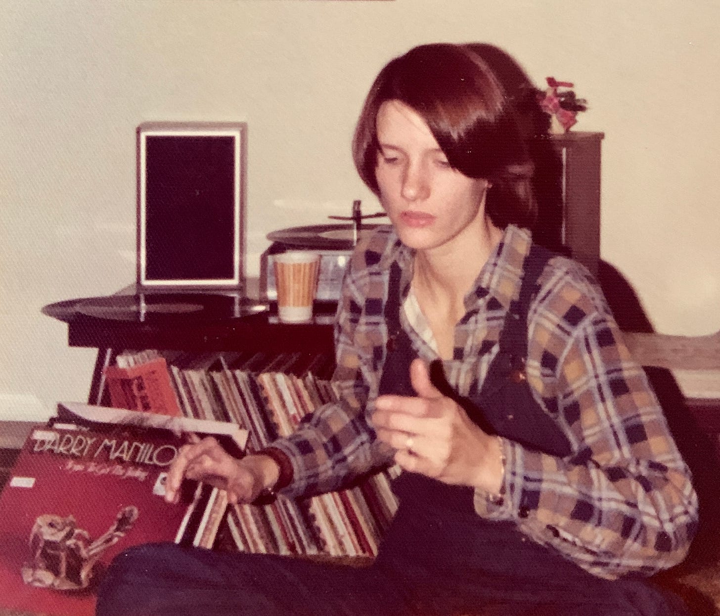A young woman in a flannel shirt and overalls with record albums all around her