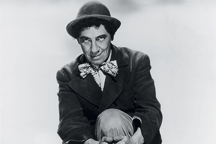 Leonard "Chico" Marx sits with his hands locked around his knee, looking up from under his little hat