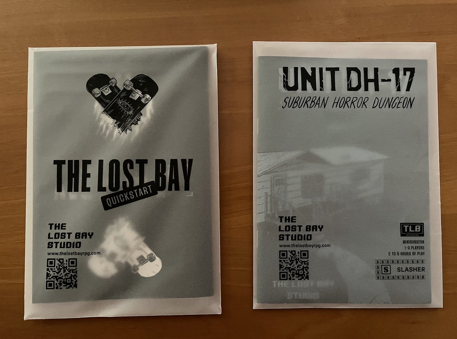 Two zines in semi-translucent vellum packaging. The covers of both zines are partially visible through the packaging, while the black text which has been printed on the vellum stands out starkly. One zine is The Lost Bay Quickstart, the other is the module Unit DH-17, both published by The Lost Bay Studio.