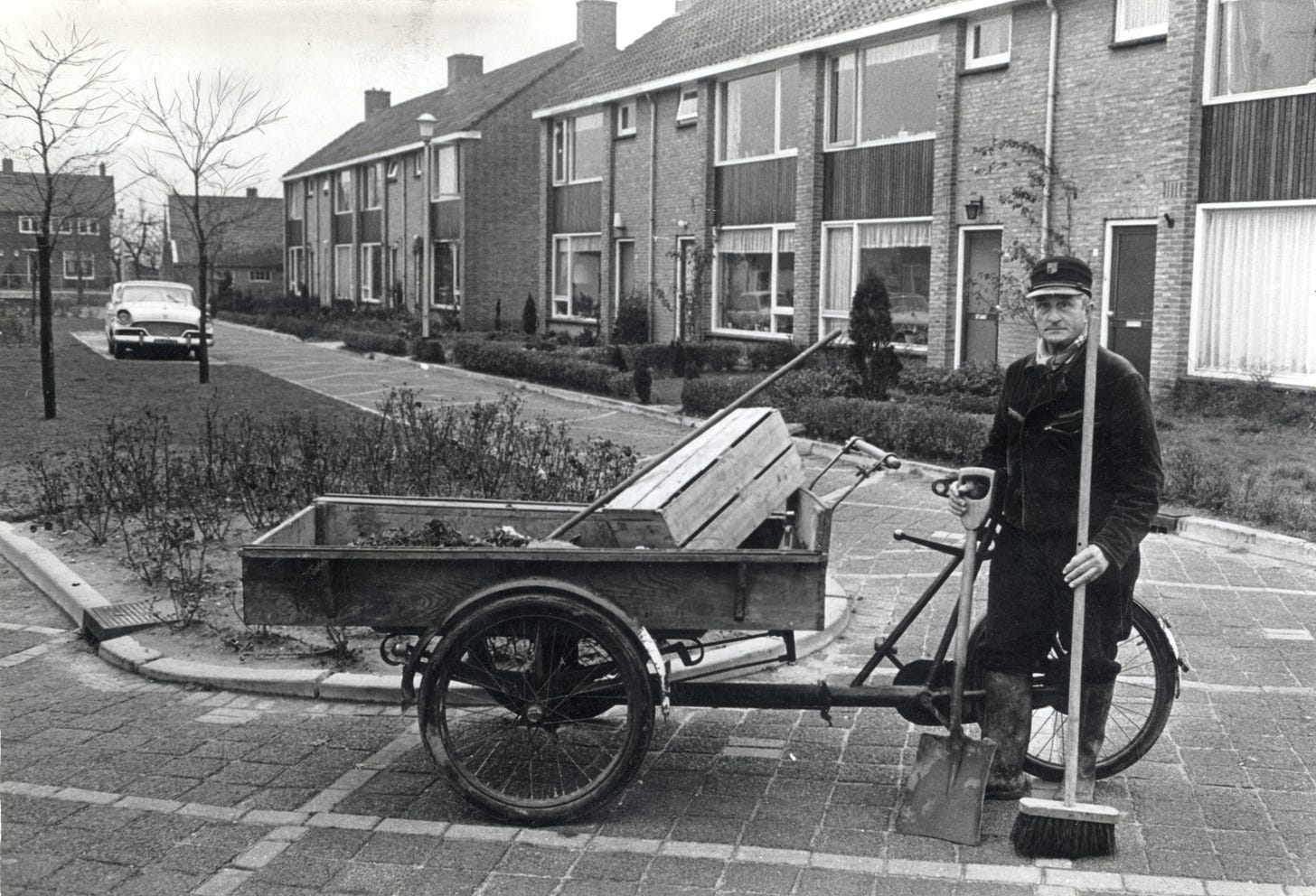 A black and white photo of a street sweeper stood next to his cargobike. He is holding a broom and a shovel. The box on the cargobike is filled with leaves and rubbish