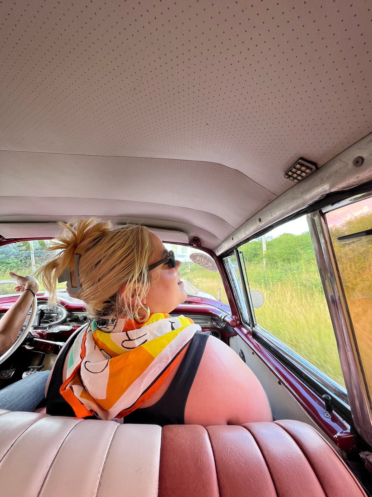 Meaghan with blonde hair and wearing sunglasses and a colourful scarf, smiling out the window of an old Cuban car