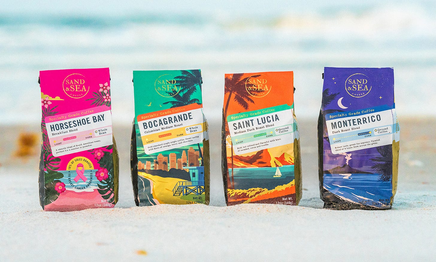 Four colorfully decorated coffee bags rest on the sand with the surf blurred in the background. The bags from left are predominantly pink, green, orange, and purple.