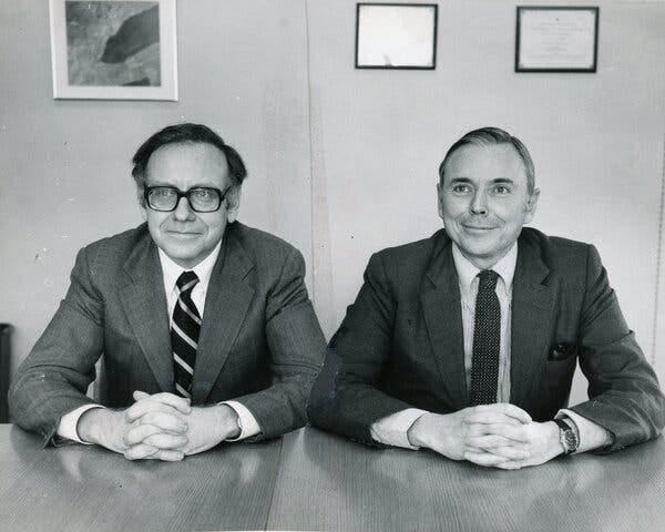 A black and white photo of the two men, both in suits and ties, sitting side by side at a table, their hands on the table folded in front of them.