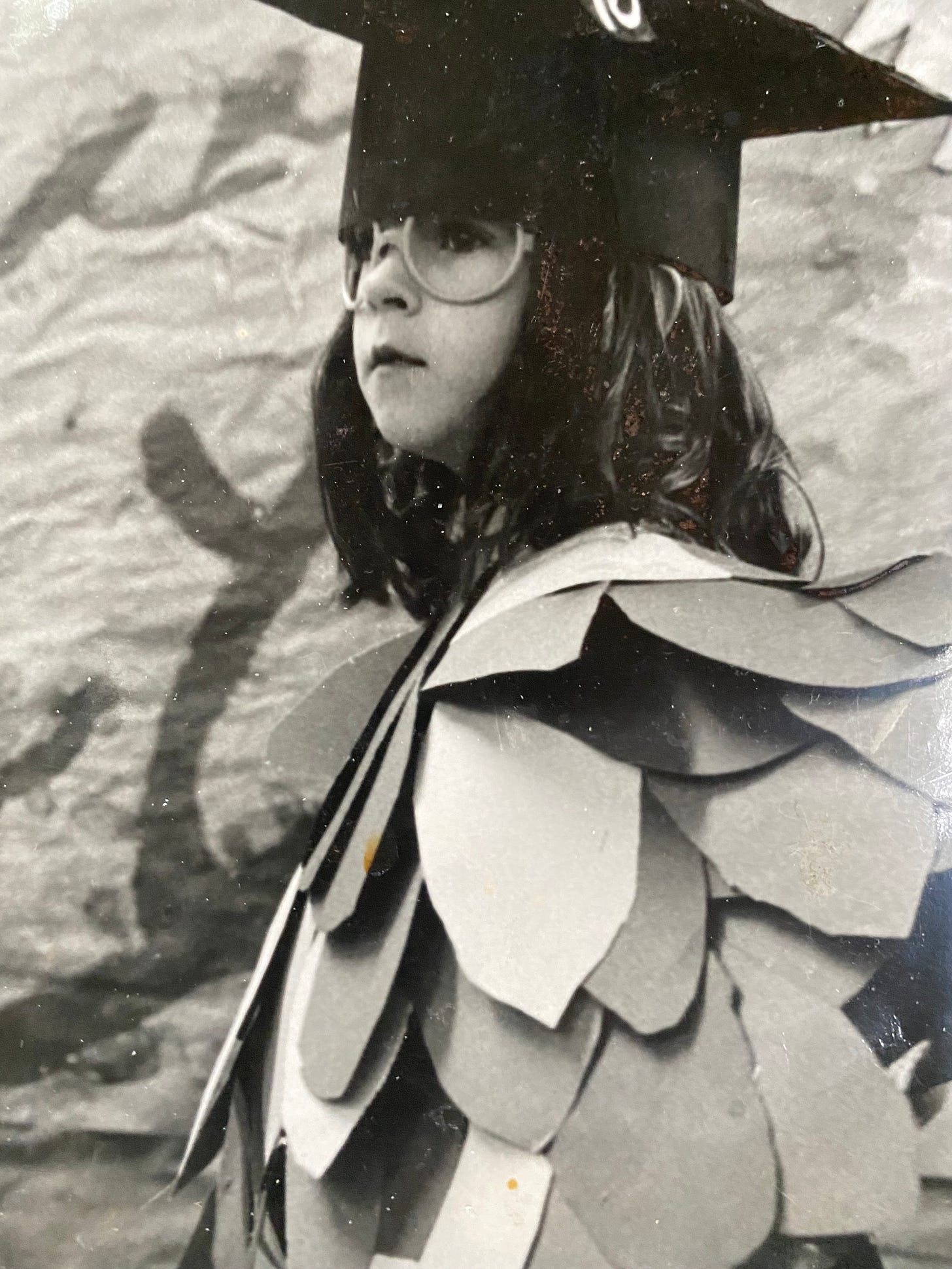 Black and white photo of the author as a child dressed in an owl costume for a school play; she is wearing a shirt of paper feathers and a black mortarboard cap with a tassel, and an intense, serious look on her face.