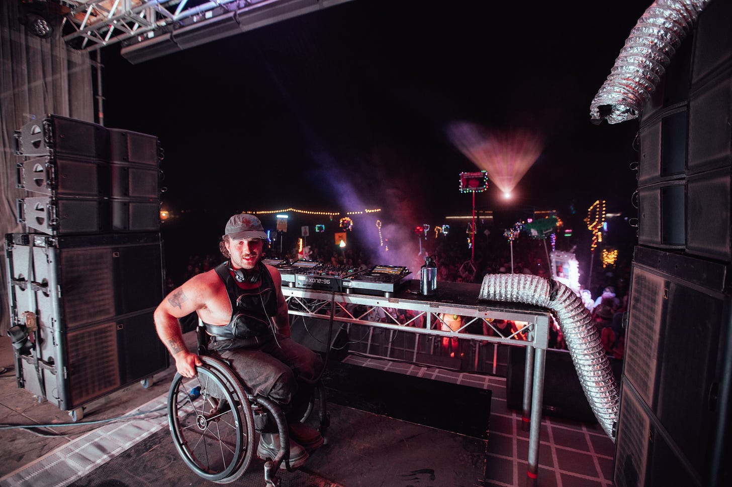 Behind the DJ booth at a large party, a white wheelchair-using man is momentarily turning away from the DJ equipment. He’s in between two walls of big speakers and subwoofers. Beyond him, there is a dark dance floor with lighted signs throughout.