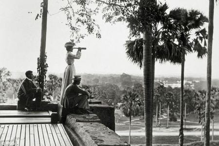 Black and white photo circa 1800s of two men and a woman. The woman is looking out aross a grove of palm trees through a spyglass.