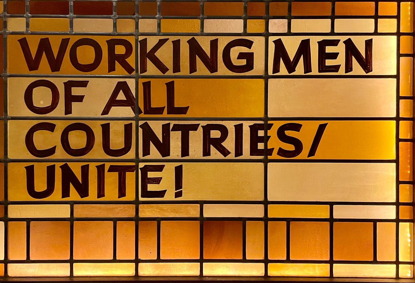 A stained glass panel in amber and ochre hues bears the bold, capitalized slogan: “WORKING MEN OF ALL COUNTRIES/UNITE!” in dark red lettering. The design is rigid and geometric, evoking Socialist-era propaganda aesthetics. A stained glass panel in amber and ochre hues bears the bold, capitalized slogan: “WORKING MEN OF ALL COUNTRIES/UNITE!” in dark red lettering. The design is rigid and geometric, evoking Socialist-era propaganda aesthetics.