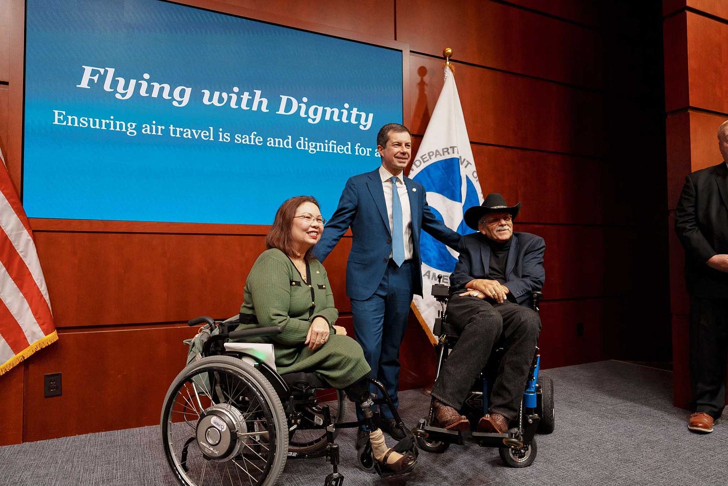 Secretary Pete standing onstage between two wheelchair users, a senator and policy advisor.