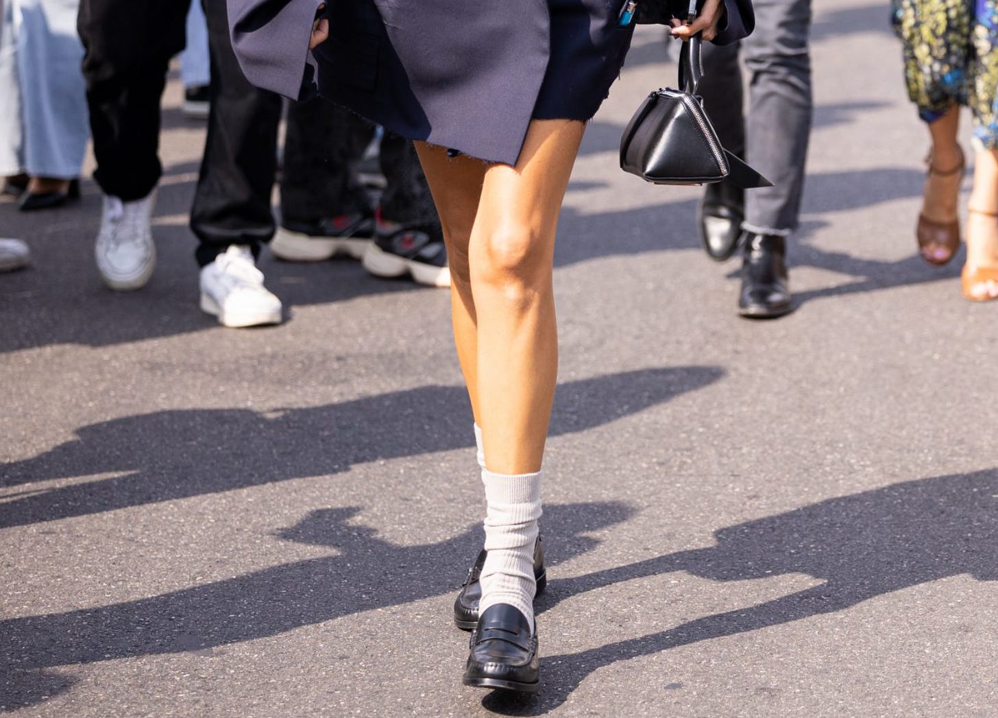 Imagem: Mulher da cintura para baixo, usando um terno oversize, pernas de fora, meia bege até o tornozelo e sapatos mocassim