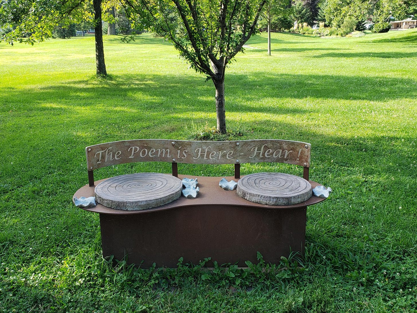 Green park setting with a sculptured metal bench that says "The Poem is Here - Hear!"