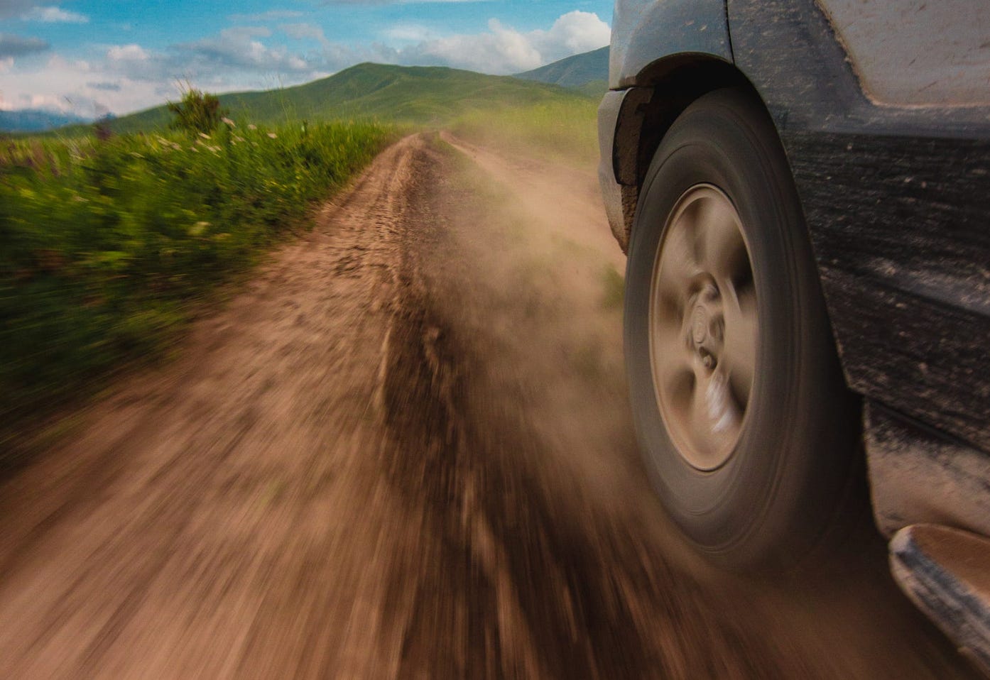 Photograph of the front wheel of a car going at speed down a dirt track. There's mud on the underside of the car. The sky is blue, and there are green hills in the background and a grassy meadow each side of the dirt road.