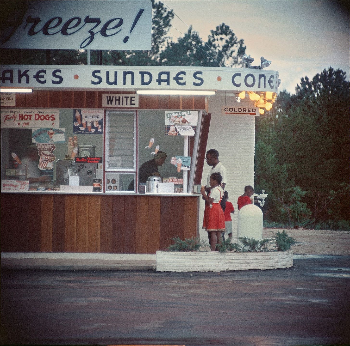 A photo taken in 1950s Alabama showing a Black father with three young children being served at an ice cream counter at a side window labelled “COLORED” by a White woman. The photographer is positioned at a distance showing is a the front of the shop and its main window labelled “WHITE” — demonstrating that this photo was taken at a time when racial segregation was still legal in the United States.